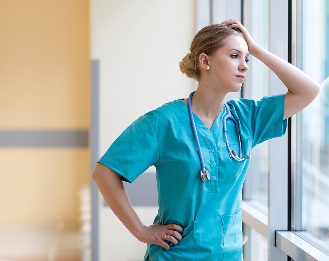 An exhausted nurse looks out the window of a healthcare facility.