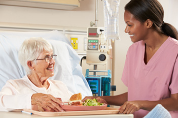 A smiling nurse watches an elderly woman in a hospital bed eating her meal from a tray.