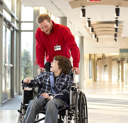 An Aramark employee in a red shirt pushing a young boy in a wheelchair with a sling on his arm.