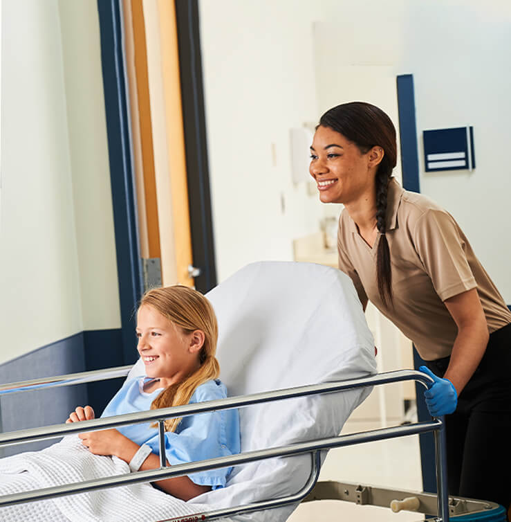 A healthcare worker pushes a wheeled hospital bed with a young patient in it.