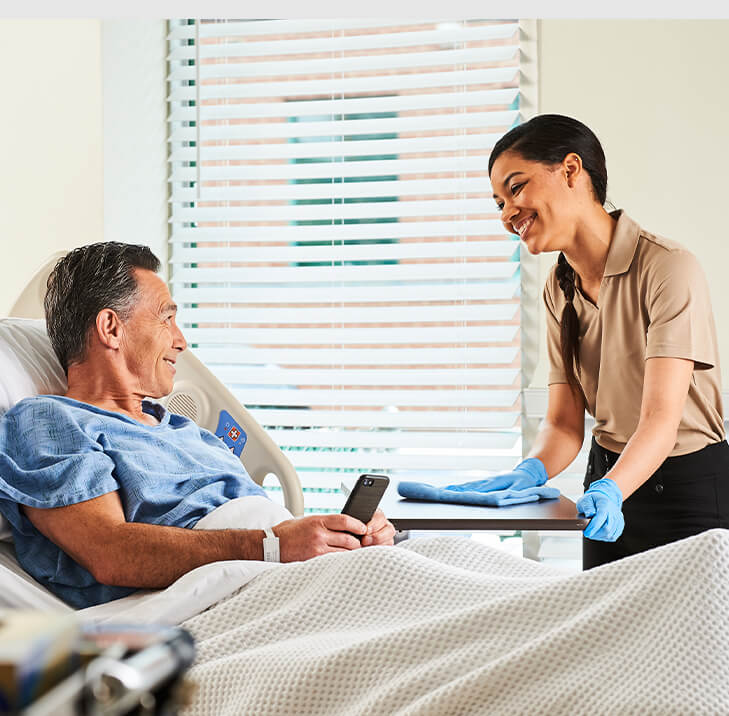 A hospital employee wipes down a bedside tray table while talking with a patient.