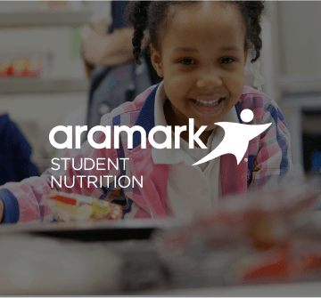 A young child holding a plate of food and smiling with the Aramark Student Nutrition logo.