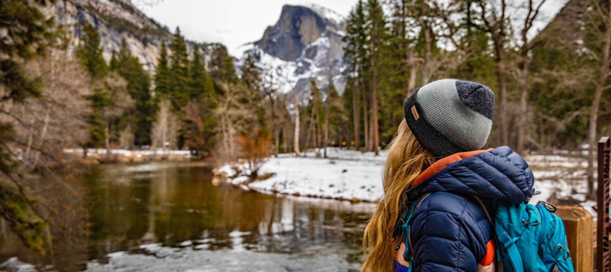 A hiker wearing a backpack looks at snow-covered mountains beyond a lake.